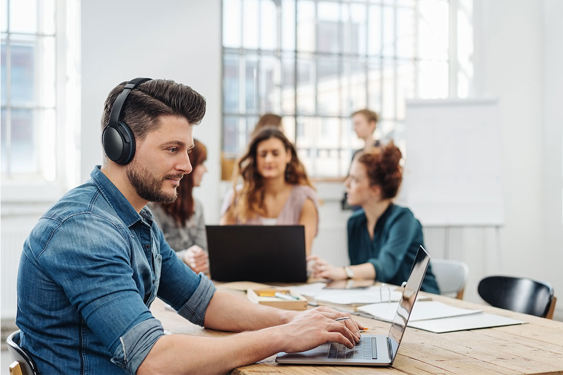 Modern office with business team working on laptops seated around a table with young man wearing headphones typing on a laptop