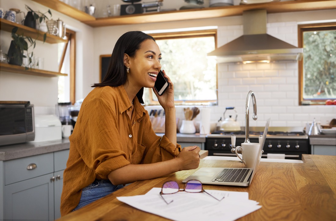 woman sitting at a table in her kitchen with her laptop on the table in front of her while she is talking on the phone