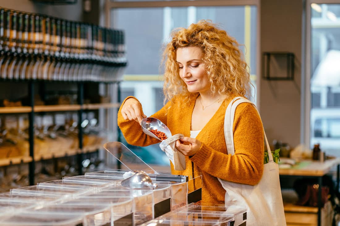 Sustainable shopping at small local businesses. Blond curly female in yellow cardigan putting Goji berries in cotton produce bag at plastic free store.